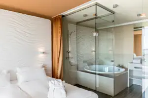 Suite bij de aja Warnemünde Een badkamer met een glazen douche, bad en glazen wand, met het bed ervoor.