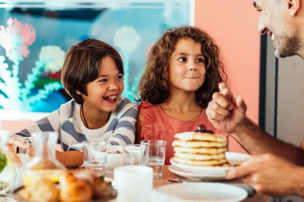 Twee kinderen zitten met hun vader in de familieruimte aan een tafel met pannenkoeken, drankjes en broodjes.