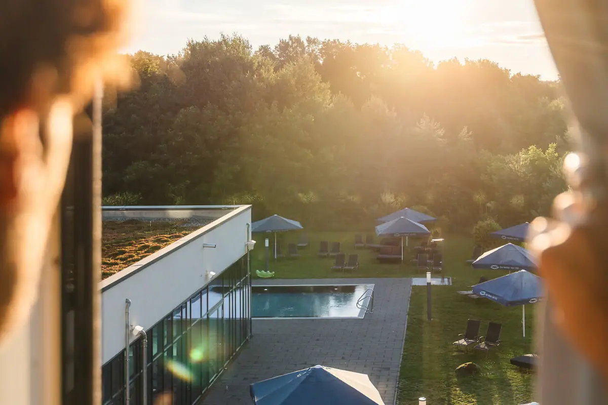 Uitzicht vanuit de kamer bij aja Bad Saarow Een persoon kijkt uit een raam naar een zwembad met parasols.