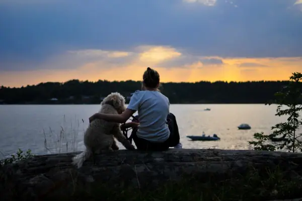 Een vrouw en een hond zitten op een boomstam bij het water.