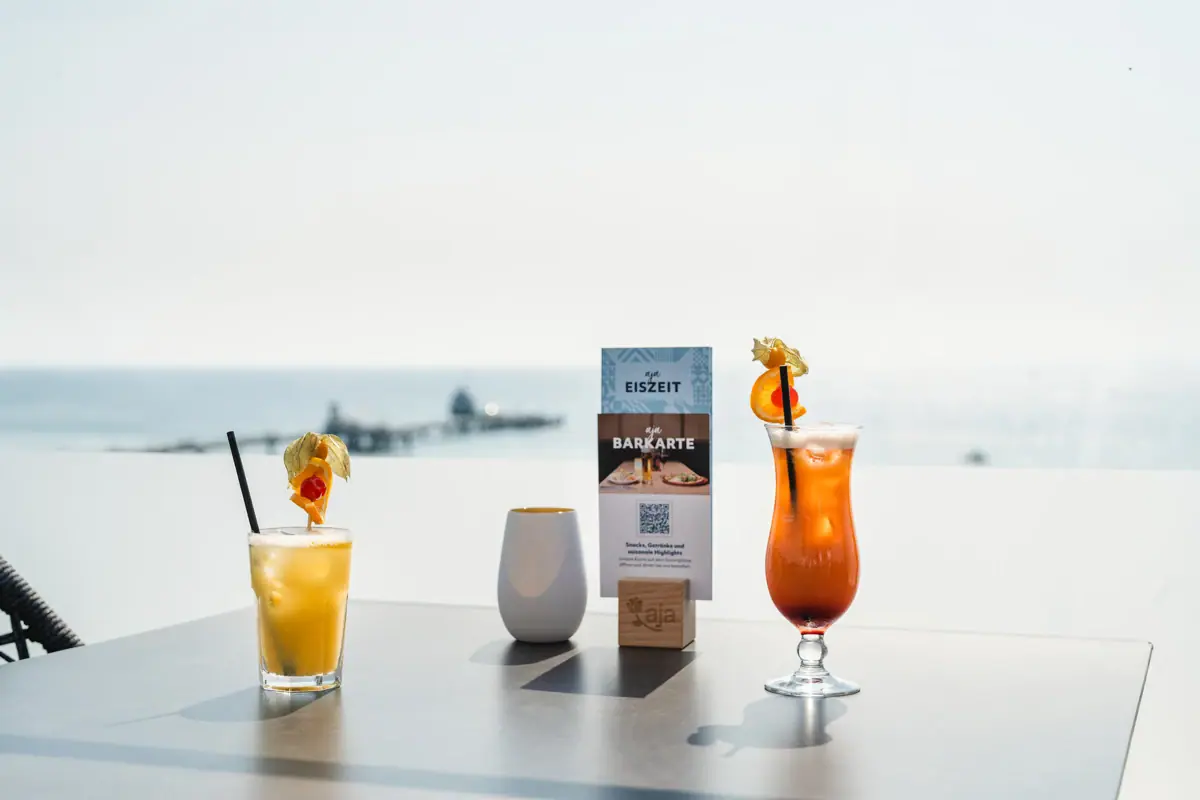 Twee cocktails op een tafel in de aja Rooftop Bar Grömitz met uitzicht op zee en de pier op de achtergrond.