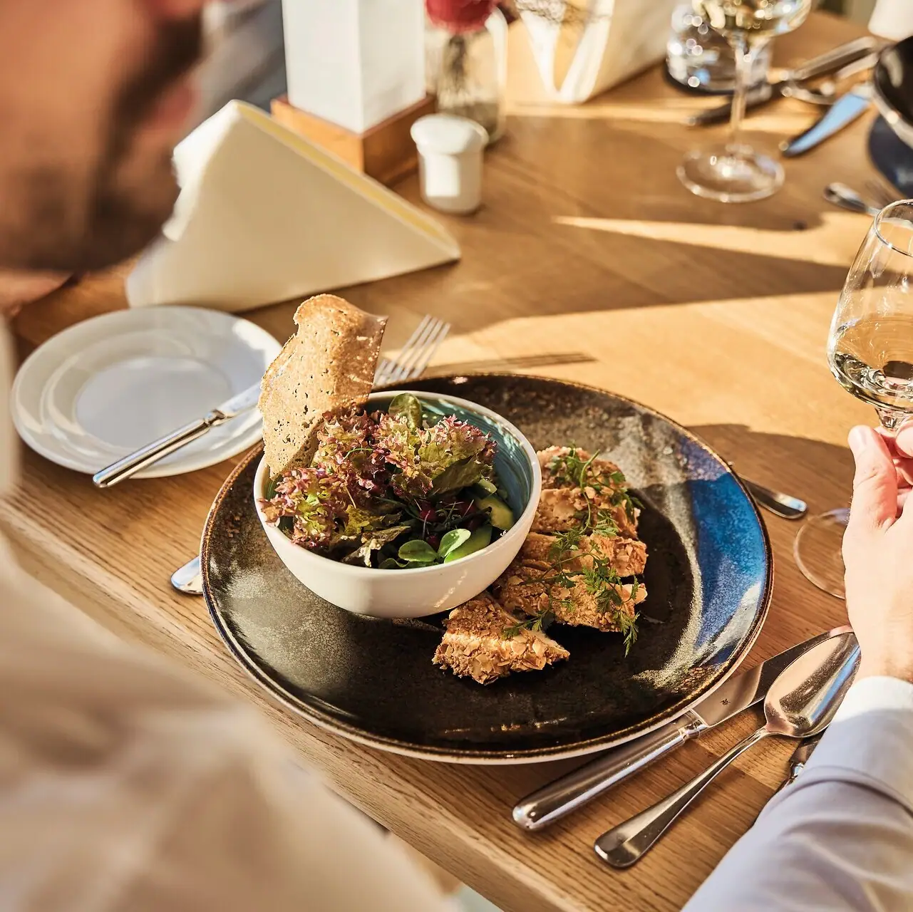 Rechtbank Een man eet van een bord en houdt een glas wijn vast. Er staan twee borden op tafel. De zon schijnt op de tafel.