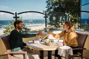 Restaurant Terrasse Ein Mann und eine Frau sitzen an einem Tisch mit Weingläsern mim Freien und im Hintergrund ist das Meer zu sehen..