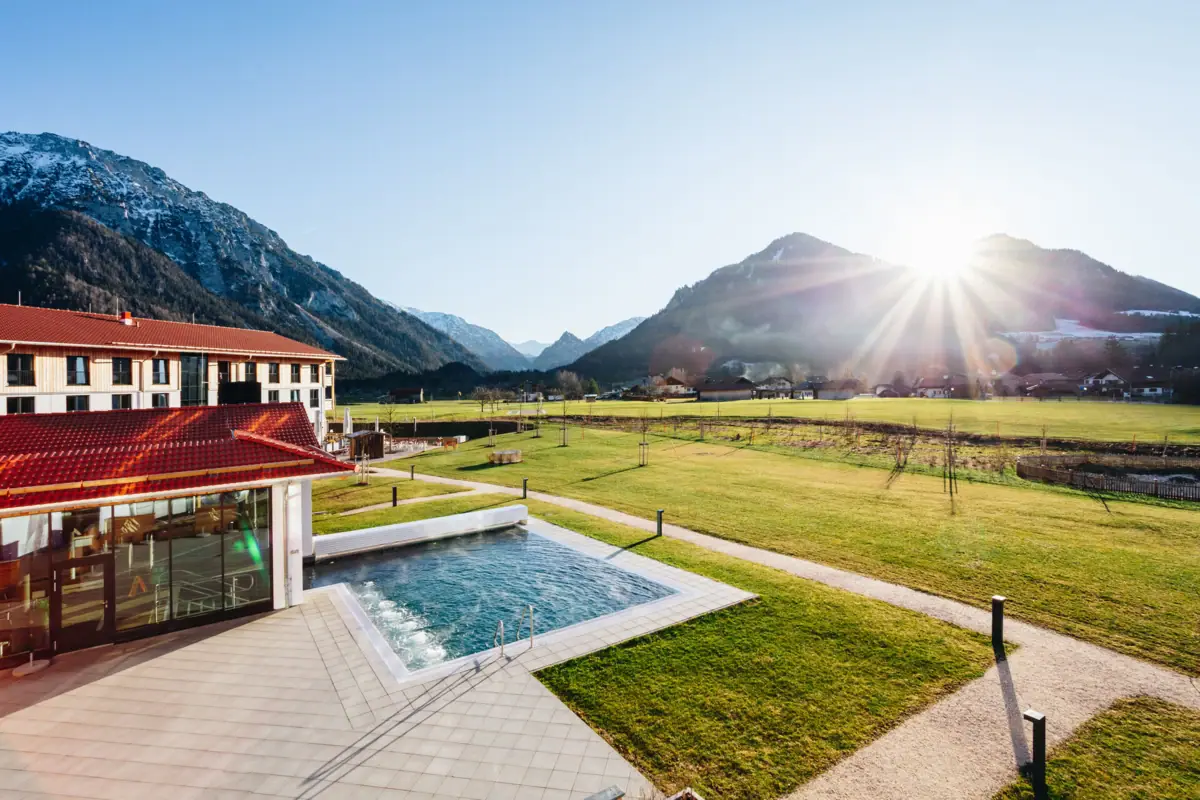 Buitenzwembad Ruhpolding Het aja Ruhpolding, een wellnesshotel in Beieren met een zwembad in een met gras begroeide omgeving, met de bergen op de achtergrond.