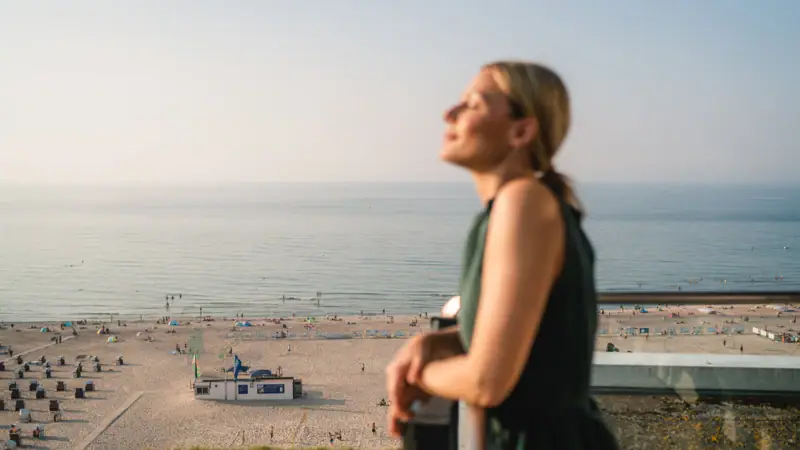 Kamers in het aja Warnemünde Een vrouw staat op een reling die uitkijkt over een strand.