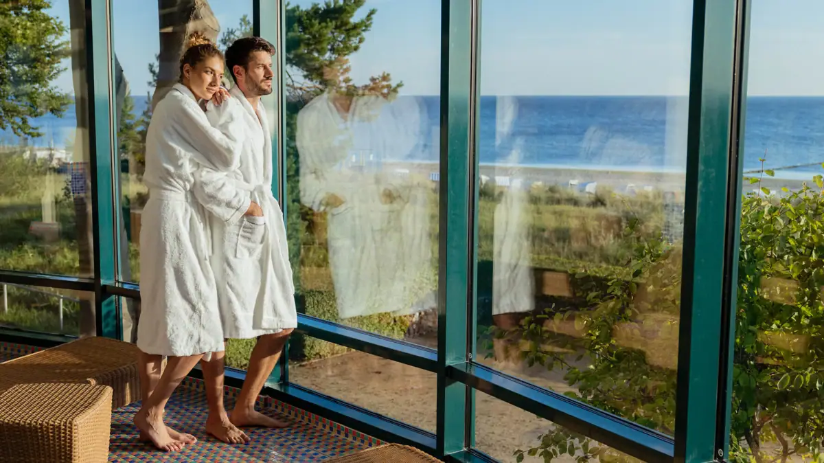 Een man en vrouw in badjassen staan in een kamer met grote ramen.