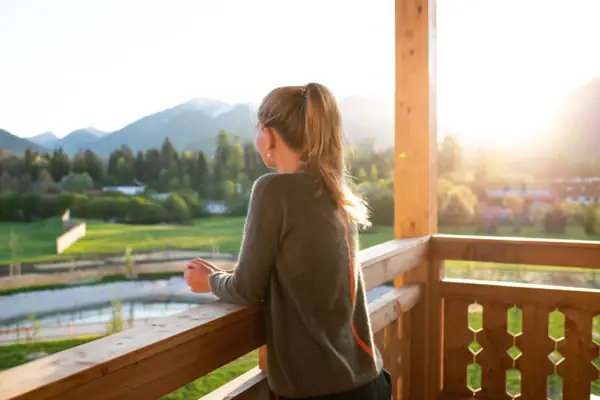 Een vrouw leunt op de houten reling van een balkon en kijkt in de verte. De ochtendzon schijnt warm in het beeld. 