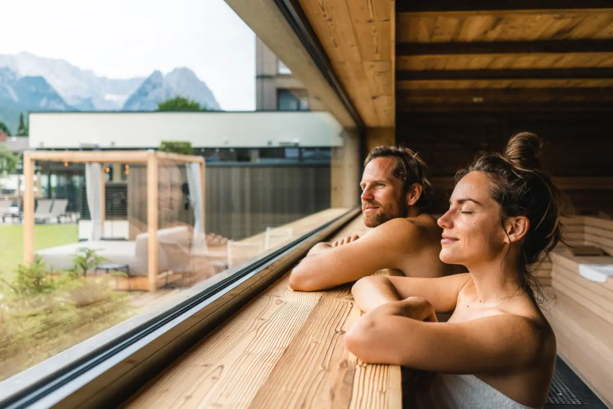 Sauna Aja in Garmisch-Partenkirchen Een man en een vrouw leunen tegen een raam in een sauna van een wellnesshotel in Beieren.