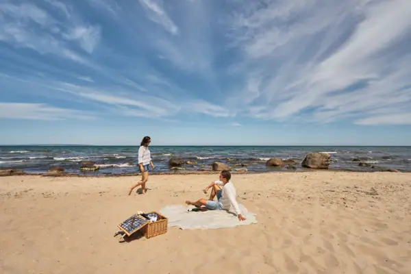 Paar aan het picknicken op een natuurlijk strand aan de Oostzee; de man zit op een deken in het zand, de vrouw loopt blootsvoets langs het water, op de voorgrond een picknickmand, op de achtergrond de zee met rotsen en een verre horizon.