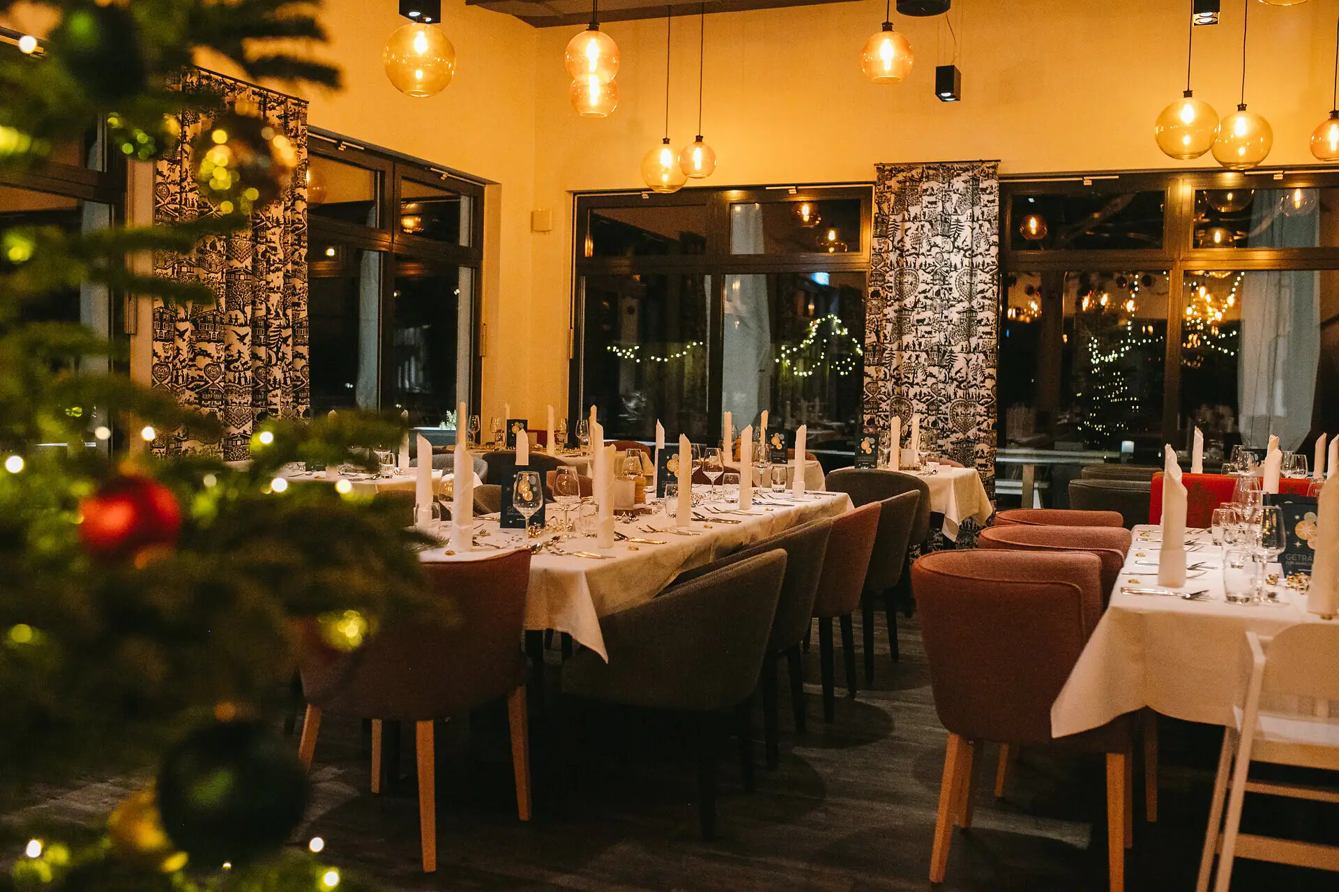Een kamer met tafels en stoelen, feestelijk versierd en een kerstboom op de voorgrond in de onscherpte.