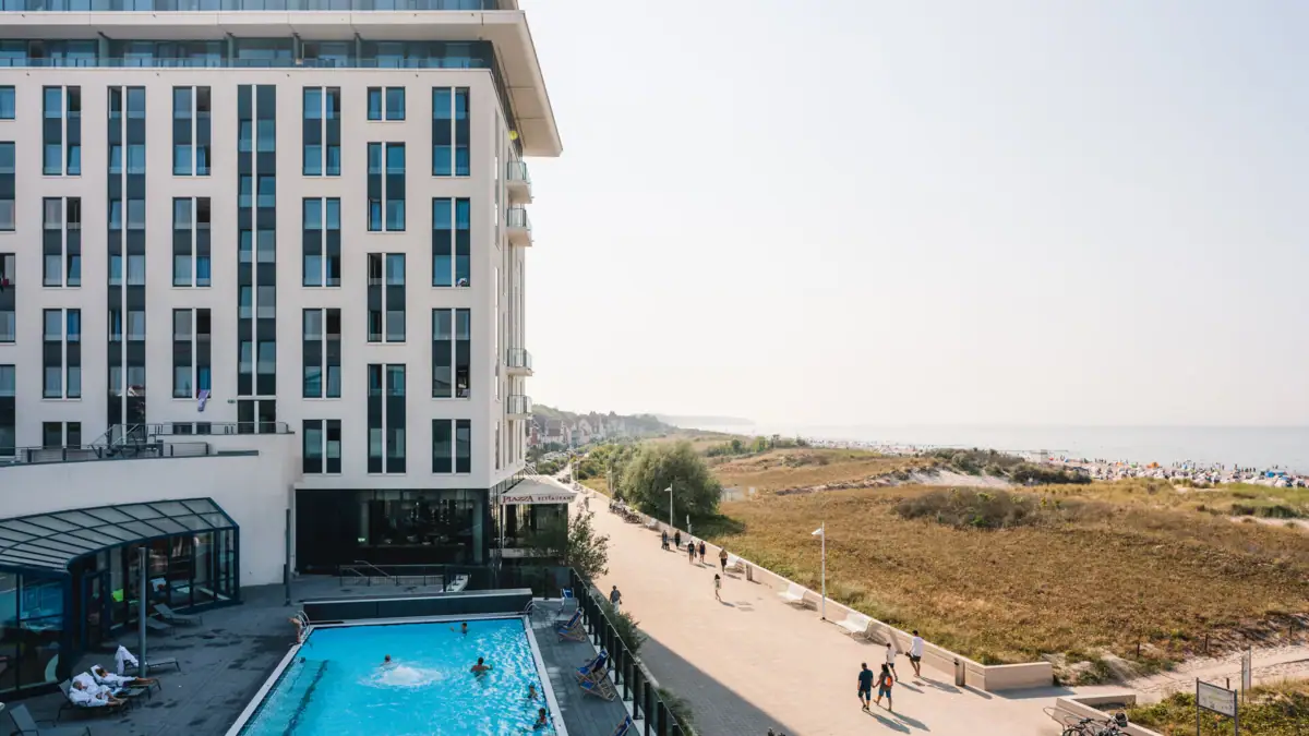 Wellness hotel aja Warnemünde Een gebouw met een zwembad, waar mensen langs lopen. De zee en het strand zijn te zien op de achtergrond.