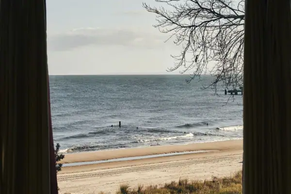 Een raam met gordijnen dat uitkijkt op een strand met zand.