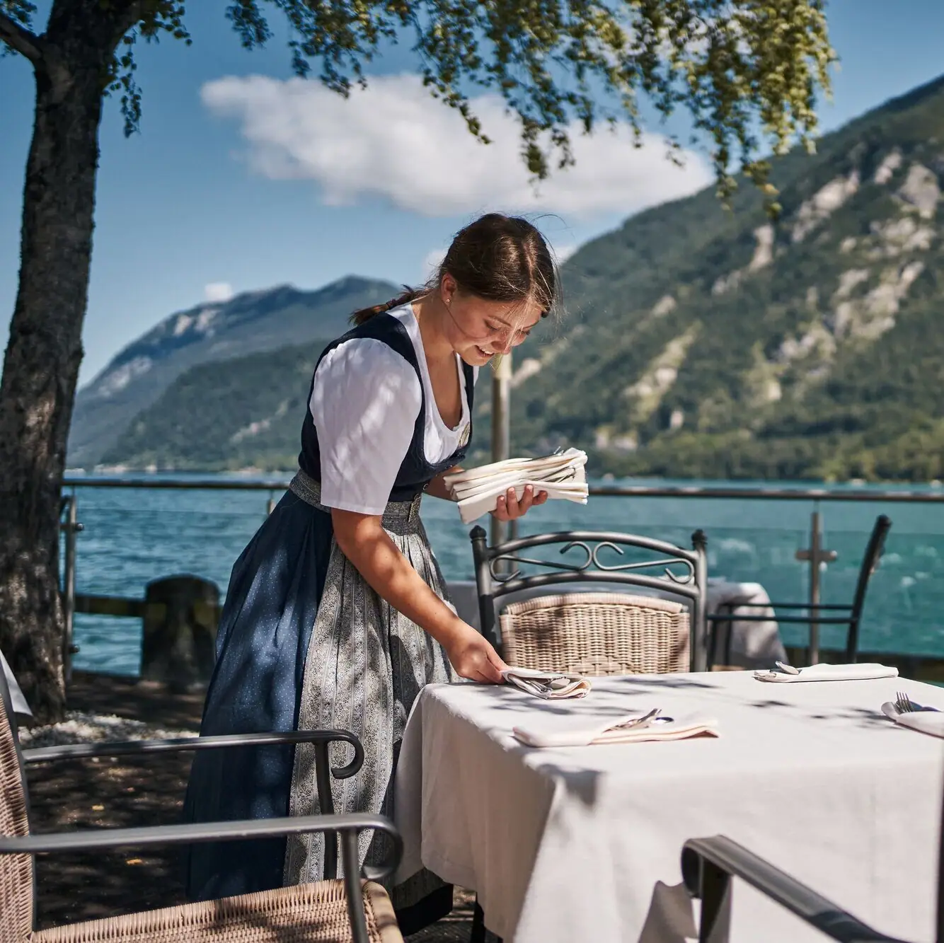 Terras aan het meer Een vrouw in een jurk aan een tafel met een meer op de achtergrond.