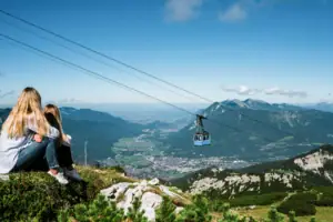 Ein Mädchen sitzt auf einem Berg und Blickt ins Tal, über ihr schwebt eine Seilbahn.