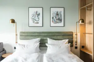 Kamers in het aja Warnemünde Een tweepersoonsbed met witte kussens en een groen hoofdeinde, geflankeerd door twee lampen en een open kledingkast