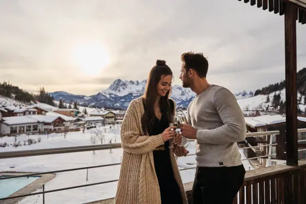Ein Mann und eine Frau stehen auf einem Balkon mit schneebedeckten Bergen im Hintergrund.