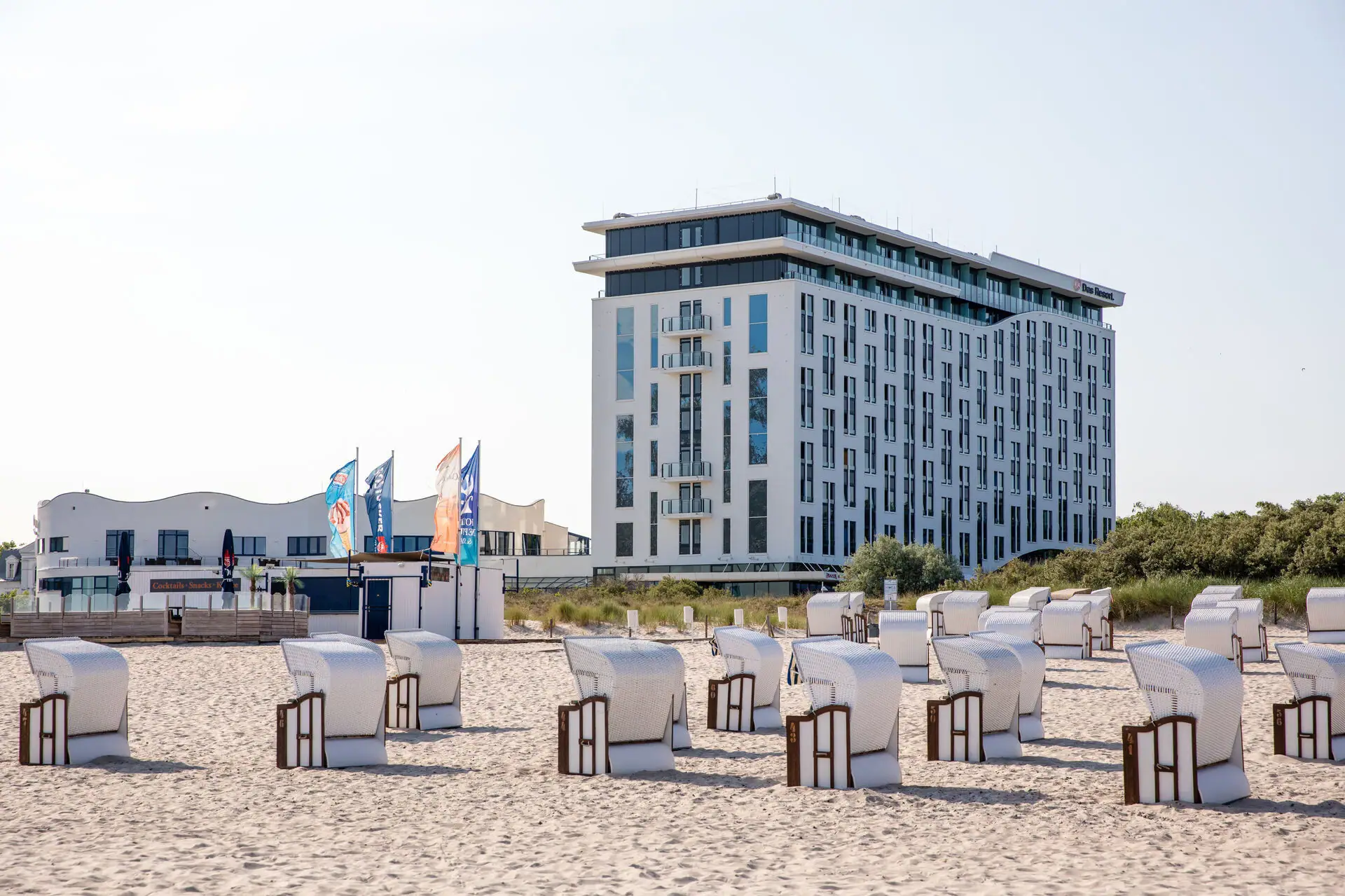 aja Warnemünde Strandstoelen staan in een rij voor een wit gebouw op het strand en de zon schijnt.