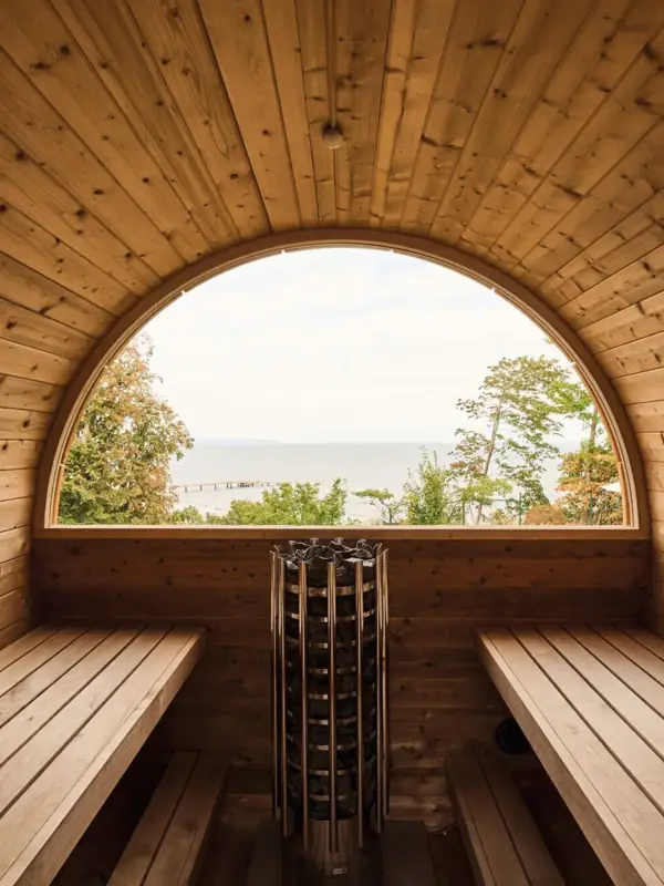Een houten barrelsauna met een rond raam dat uitkijkt over het water.