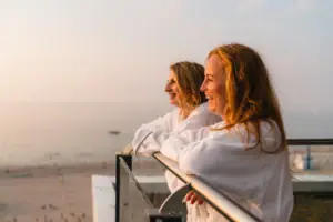 Zwei Frauen in Bademänteln lehnen an einem Geländer mit Blick auf einen Strand.
