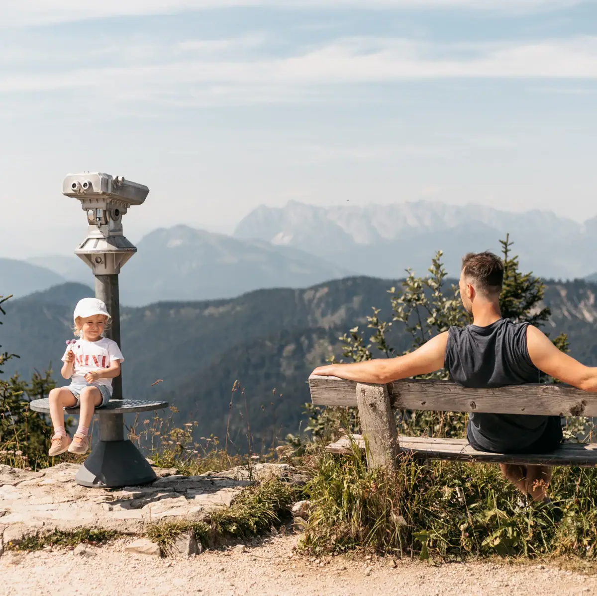 Vader en dochter genieten van het uitzicht op de Alpen vanaf de avonturenberg Rosnerköpfl in Werfenweng. Het meisje zit op een uitkijktoren, de man ontspant op een houten bank met uitzicht op het bergpanorama.