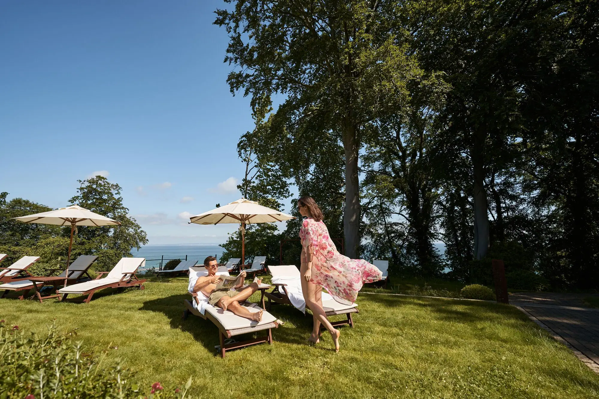 SPA ligweide Paar ontspant op ligstoelen in de tuin van een hotel met uitzicht op de Baltische Zee; de vrouw draagt een luchtige zomerjurk, de man proost met een drankje - parasols en groene bomen op de achtergrond.