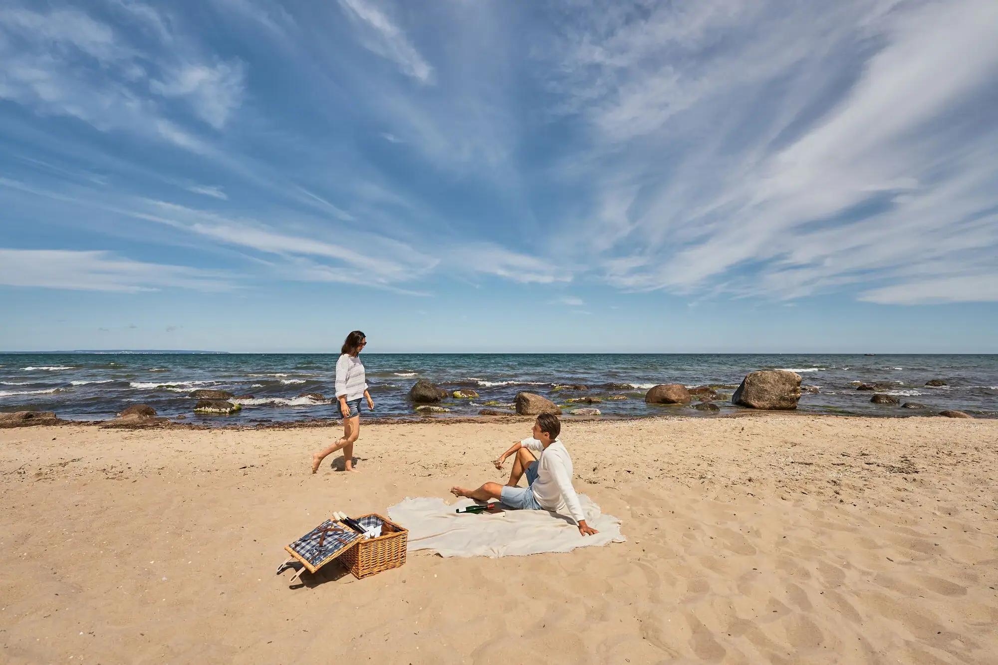 Göhren strand Paar aan het picknicken op een natuurlijk strand aan de Oostzee; de man zit op een deken in het zand, de vrouw loopt blootsvoets langs het water, op de voorgrond een picknickmand, op de achtergrond de zee met rotsen en een verre horizon.