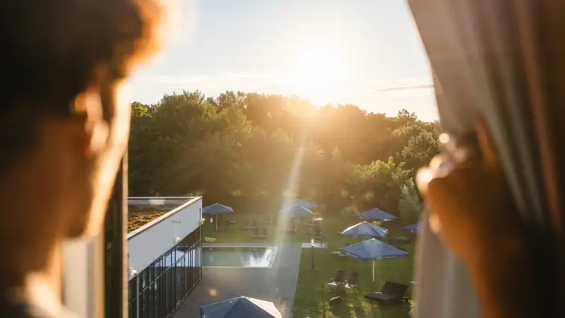 Een persoon kijkt naar een zwembad met parasols en bomen.