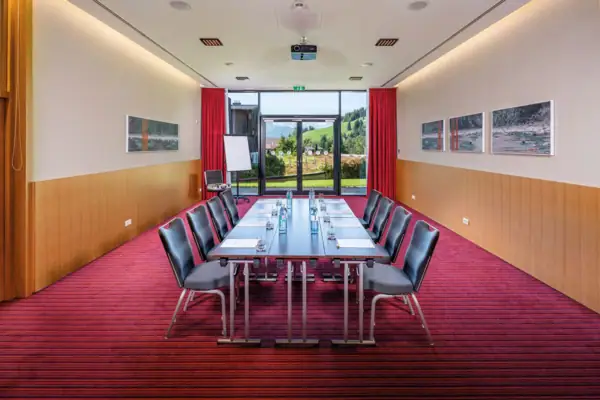 Er staat een lange tafel in een vergaderzaal met een rode loper. Daglicht valt naar binnen door een grote patiodeur aan de achterkant. 