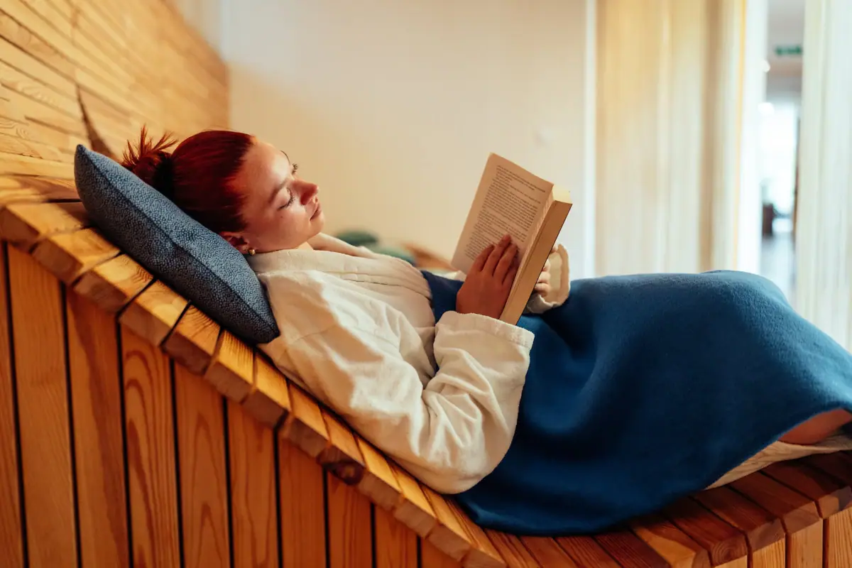 Een vrouw ligt op een houten bankje en leest een boek.