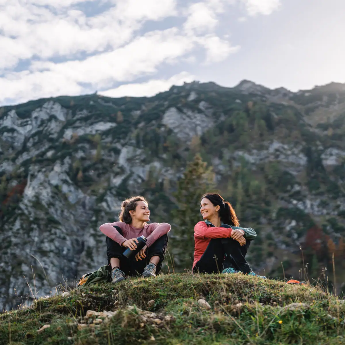 Wandelvakantie Twee vrouwen zitten op een heuvel met bergen op de achtergrond.