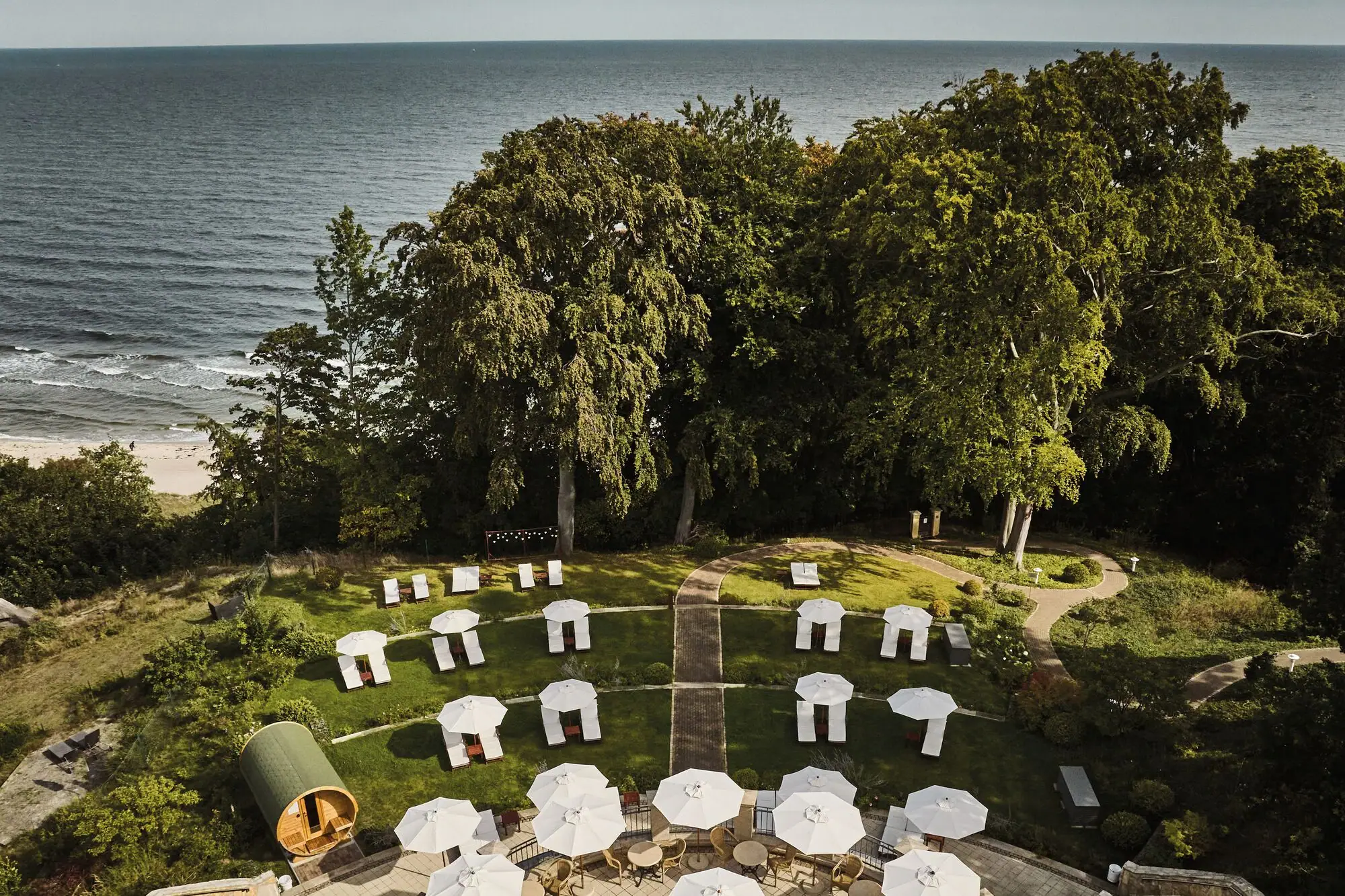 Eine Gruppe von weißen Liegestühlen und Sonnenschirmen auf einer grasbewachsenen Fläche mit Bäumen und Wasser.