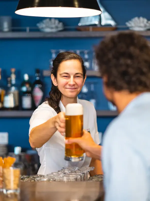 Eine Frau hält ein Glas Bier an der Bar und übergibt es einem Gast in der aja Strandbar Warnemünde.