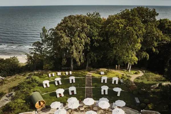 Eine Gruppe von weißen Liegestühlen und Sonnenschirmen auf einer grasbewachsenen Fläche mit Bäumen und Wasser.