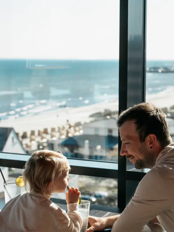 Een man en een kind zitten aan een tafel en kijken uit over een strand.