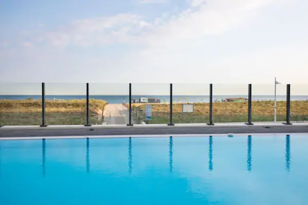 Een zwembad met een hek en gazon op de voorgrond, met daarachter een strand.