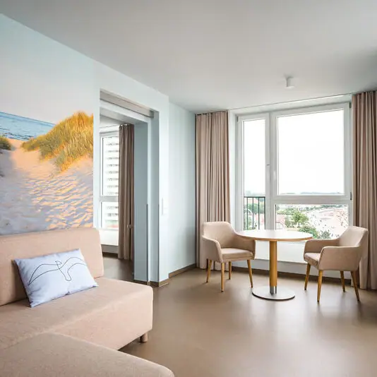 Het woongedeelte van een hotelkamer, bestaande uit een lichtgekleurde bank en twee stoelen met een ronde tafel voor een raam van vloer tot plafond. Voor het raam hangen lange beige gordijnen en achter de bank hangt een grote foto van duinen, strand en zee.