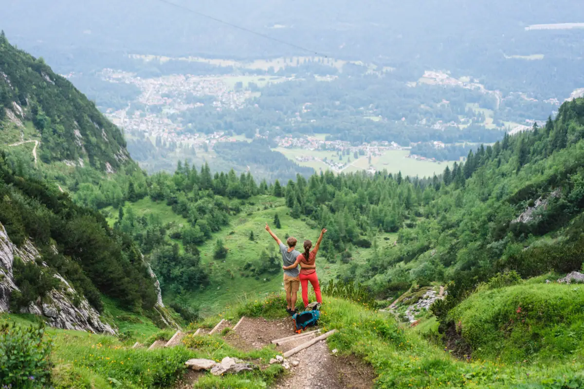 Wandelen Garmisch-Partenkirchen Een man en een vrouw staan tijdens een wandeling op een berg en kijken naar beneden in de vallei, terwijl ze hun armen in de lucht strekken.