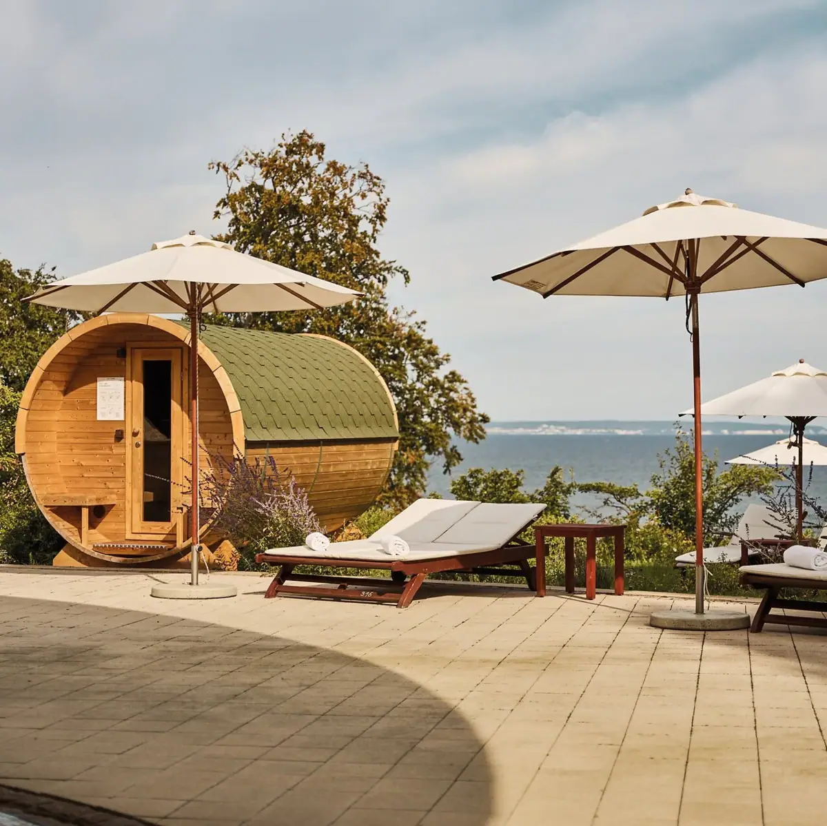 Barrelsauna met uitzicht op zee Ligstoelen en parasols naast een ton en een waterpartij.