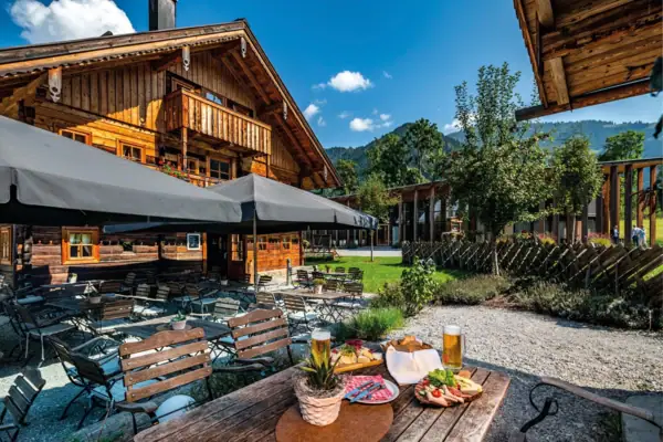 Gezellige biertuin voor een traditionele alpenhut met rustieke charme in Werfenweng. Houten tafels en stoelen onder grote parasols nodigen uit om te blijven hangen. Een bierglas, een snackschaal met worst, kaas en verse groenten en een gedekte tafel staan op de voorgrond.