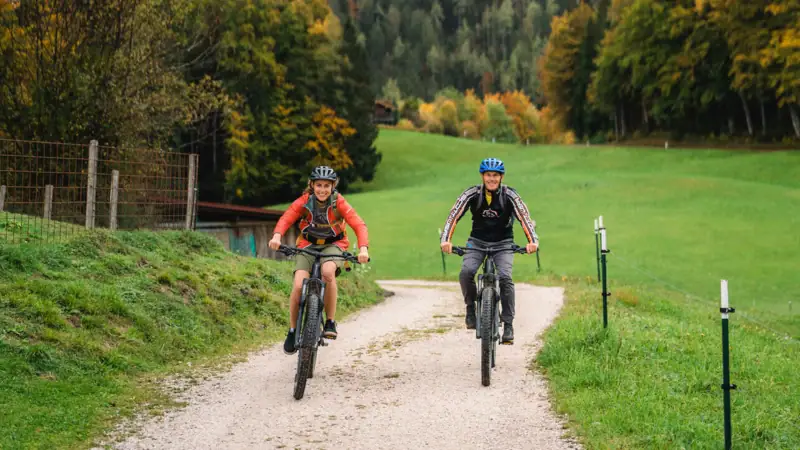Fietstocht Twee mensen fietsen op een zandpad, met bomen en bergen op de achtergrond.