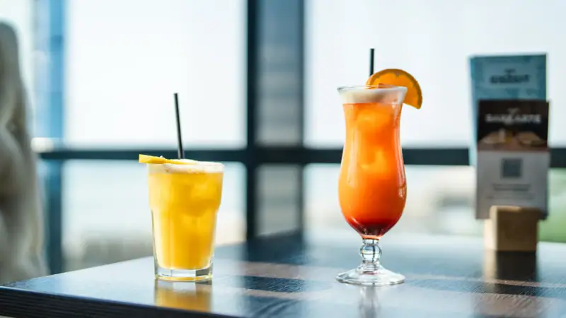 Twee glazen met drankjes op een tafel, een met sinaasappelsap en de andere met een cocktailgarnituur.