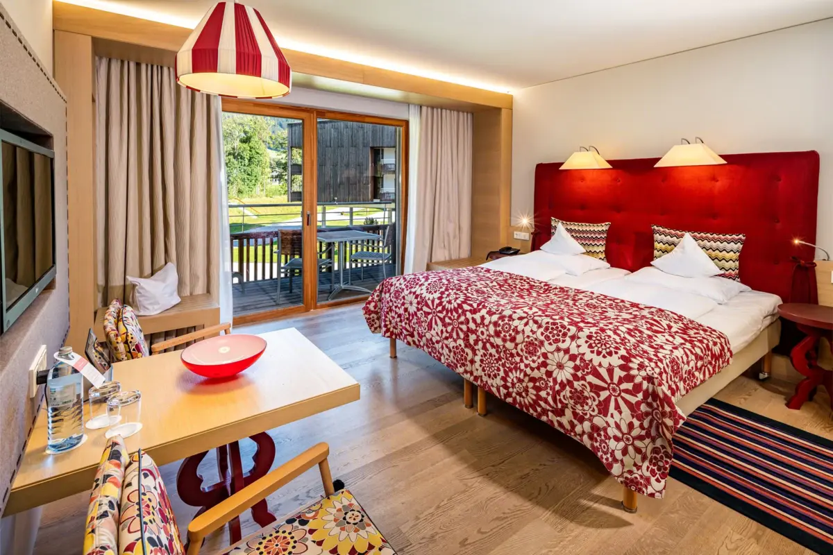 Lichte en kleurrijke hotelkamer in alpine stijl met tweepersoonsbed, rood hoofdeinde, sprei met patroon, zithoek en toegang tot het balkon met uitzicht op het groen.