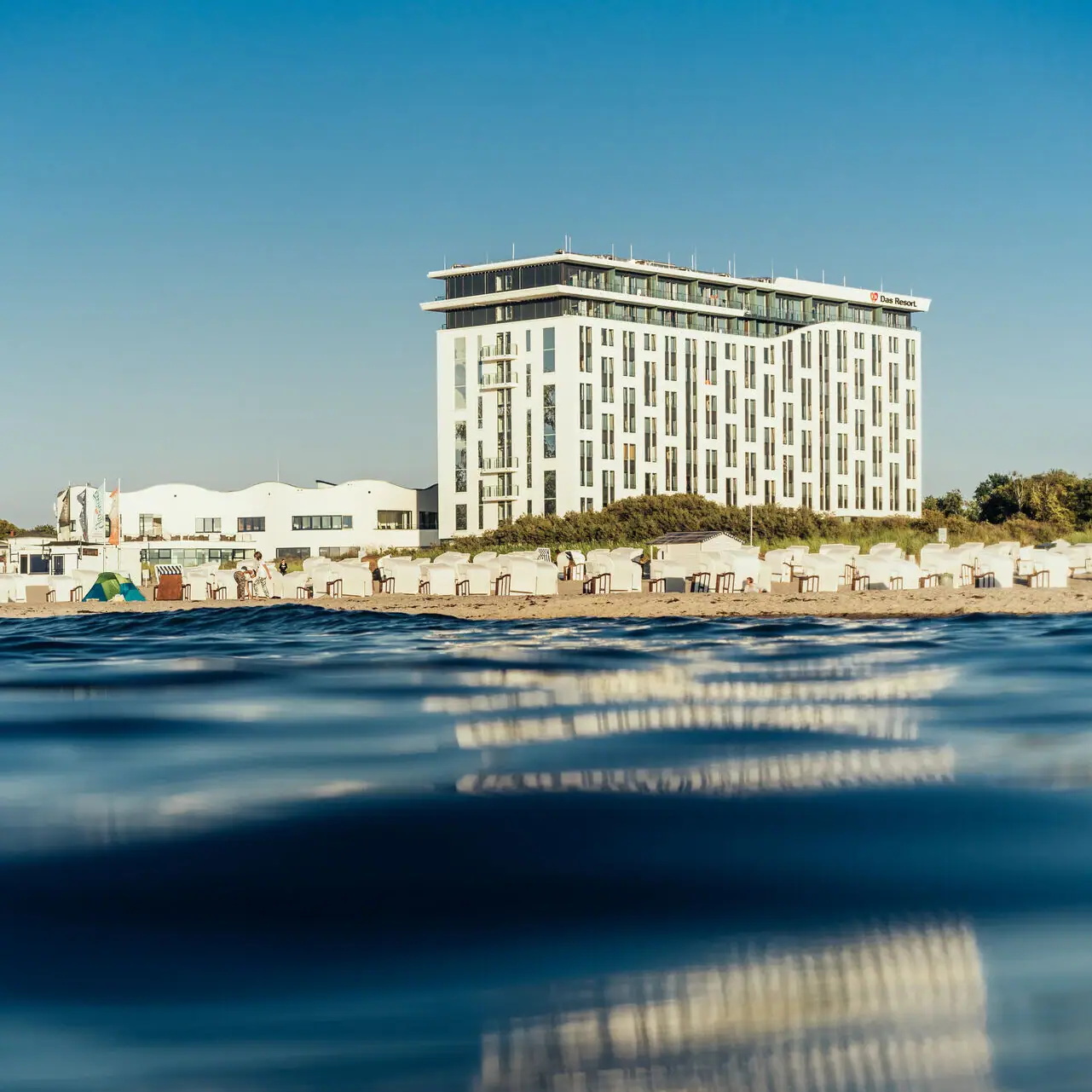 Het hotelgebouw op het strand met een heldere lucht en water op de voorgrond.