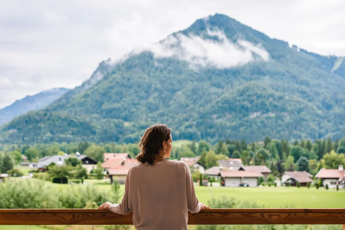 Uitzicht vanuit het aja Ruhpolding Een vrouw staat op een balkon en kijkt uit over een bergketen.