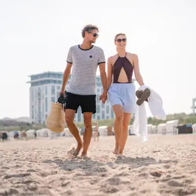 Een man en een vrouw houden elkaars hand vast op het strand, met de aja Warnemünde op de achtergrond.
