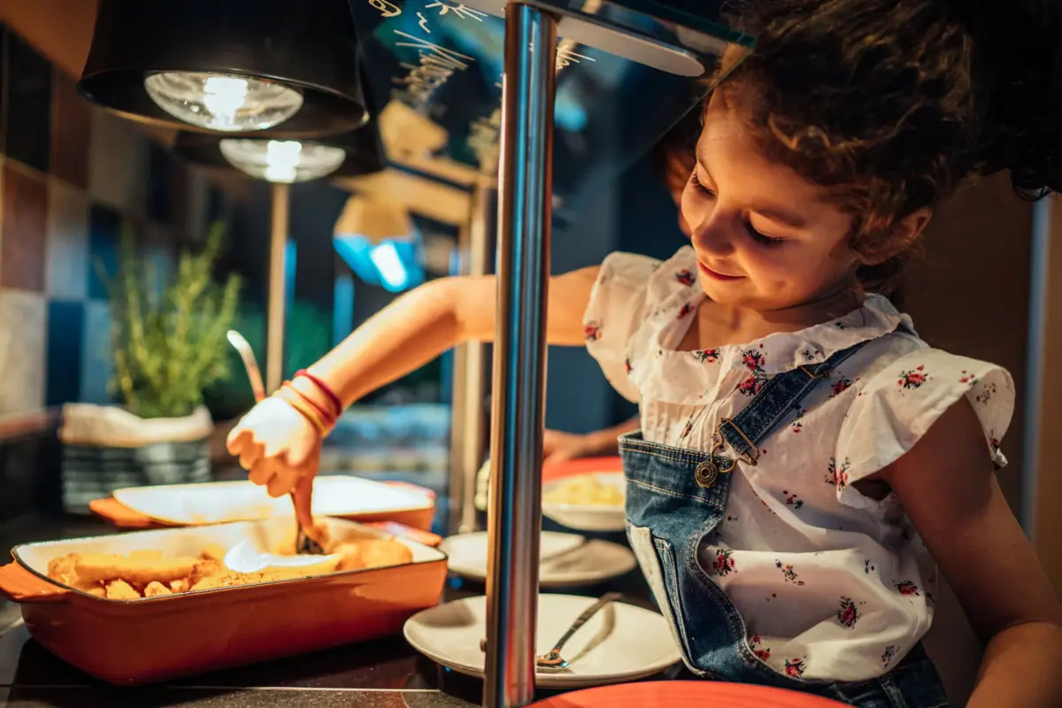 Een meisje in een tuinbroek en een wit overhemd staat bij het aja-kinderbuffet.